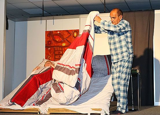 Ein erwachsener Mann in karierten Pyjamas hält eine Decke und scheint das Bett zu reparieren, während ein anderer Mann darauf liegt. Das Bett steht auf einer Holzplattform.