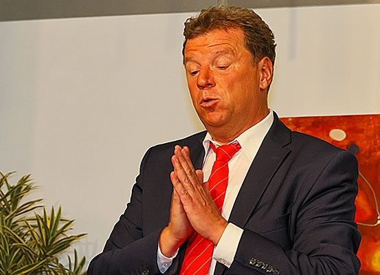 Ein Mann im Anzug klatscht und steht vor einer weißen Wand mit einer Pflanze.