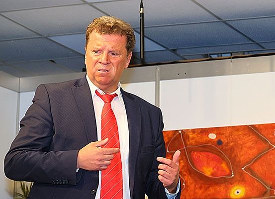 Ein Mann in Anzug und roter Krawatte hält eine Rede mit Handgesten in einem Büroraum mit einem Gemälde im Hintergrund.