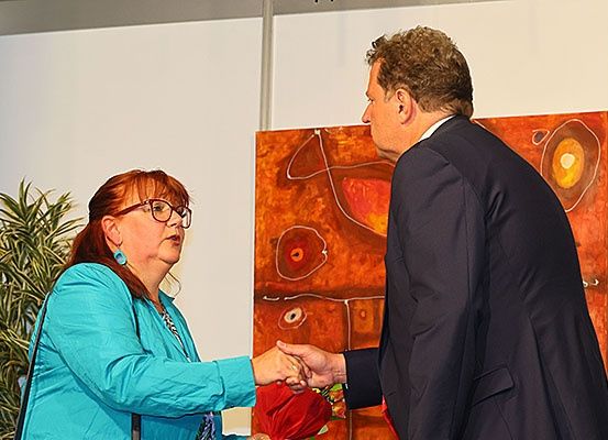 Eine Frau in einer türkisfarbenen Jacke und ein Mann in einem schwarzen Anzug schütteln sich die Hände vor einem großen orangefarbenen abstrakten Gemälde.