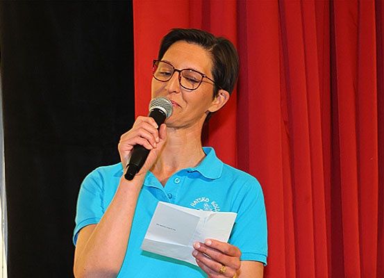Eine Frau in einem blauen Shirt liest von einem Stück Papier, während sie ein Mikrofon hält.