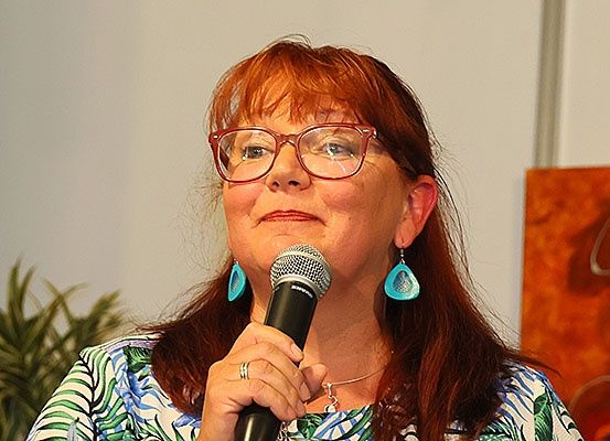Eine Frau mit langen roten Haaren und Brille hält ein Mikrofon, wahrscheinlich bei einer Veranstaltung. Sie trägt ein Blumenhemd und Ohrringe. Hinter ihr ist eine Pflanze und eine Wand zu sehen.