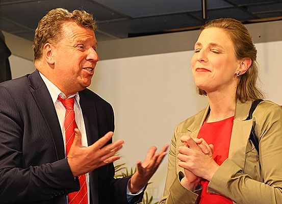 Ein Mann in Anzug und eine Frau in rotem Hemd unterhalten sich mit Handgesten.