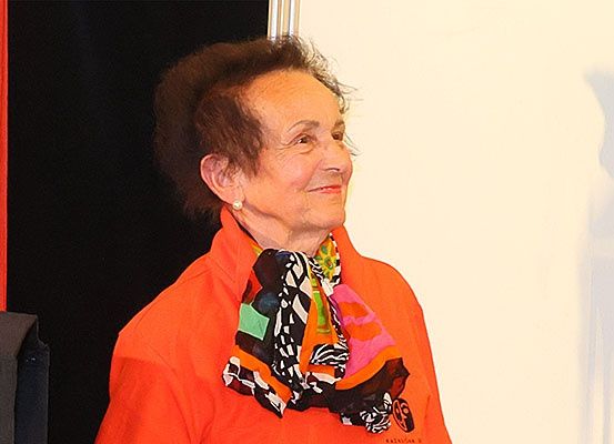 Eine ältere Frau mit kurzen Haaren lächelt und steht vor einer weißen Wand, trägt eine orangefarbene Jacke und einen gemusterten Schal.