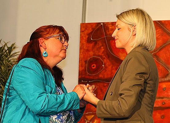 Zwei Frauen stehen vor einem großen abstrakten Gemälde, halten Händchen und lächeln sich an.