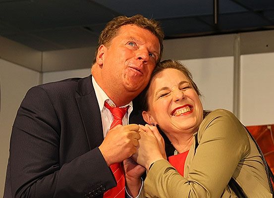 Ein Mann und eine Frau, beide in formeller Kleidung, posieren für ein Foto und zeigen übertriebene Ausdrücke. Der Mann hält die Hand der Frau und sie lehnt sich an ihn.