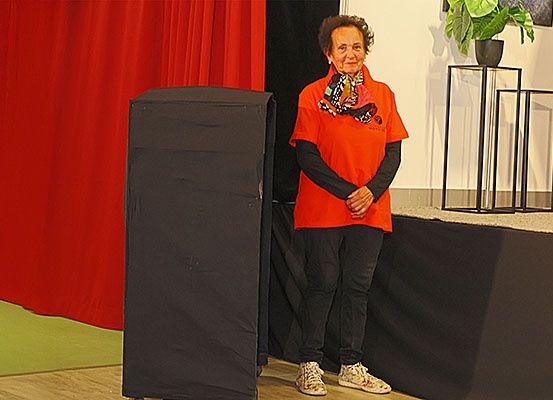 Eine ältere Frau steht auf einer Bühne mit schwarzem Hintergrund, trägt ein orangefarbenes Shirt und schwarze Hose. Sie lächelt und hat die Hände vor sich verschränkt.
