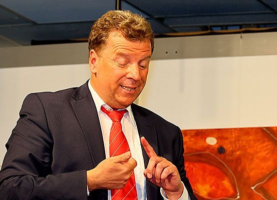 Ein Mann in Anzug und Krawatte steht in einem Raum, macht Handbewegungen und lächelt in die Kamera.