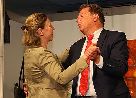 Ein Mann und eine Frau tanzen, beide lächeln. Der Mann trägt eine rote Krawatte und einen schwarzen Anzug, während die Frau einen hellbraunen Blazer und eine schwarze Tasche hat. Hinter ihnen ist eine weiße Wand mit einem Gemälde.