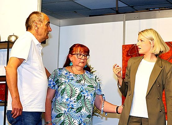 Drei Personen stehen in einem Raum mit hellem Hintergrund. Ein Mann und eine Frau stehen sich gegenüber, während eine Frau in der Mitte den Mann ansieht.