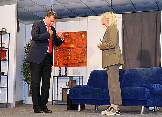 Ein Mann und eine Frau in formeller Kleidung stehen in einer Innenumgebung, wahrscheinlich auf einer Bühne. Der Mann gestikuliert, während er spricht. Die Frau steht mit gefalteten Händen vor sich. Hinter ihnen befindet sich eine blaue Couch, ein Gemälde und ein Vorhang.