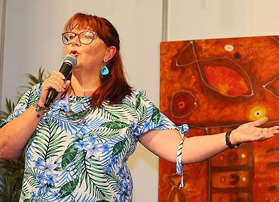 Eine Frau spricht in ein Mikrofon vor einem abstrakten Gemälde, trägt eine Brille und ein Blumenhemd.