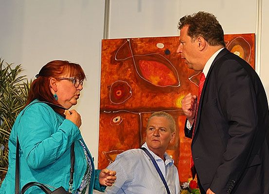 Drei Personen stehen vor einem lebhaften orangefarbenen Gemälde. Ein Mann in einem schwarzen Anzug und roter Krawatte sieht den anderen beiden gegenüber, die sich zu unterhalten scheinen.