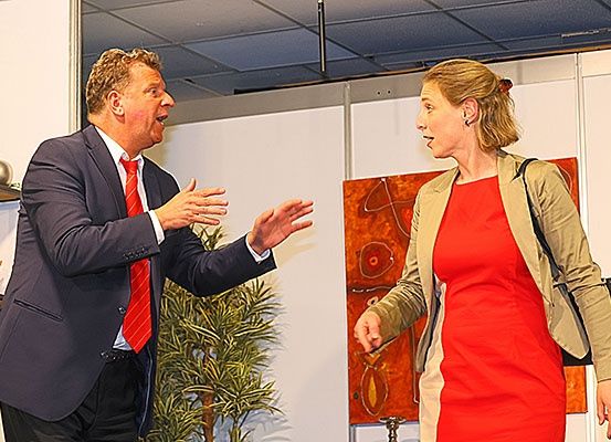 Ein Mann in Anzug und roter Krawatte spricht lebhaft zu einer Frau in einem roten Kleid, die eine Handtasche trägt. Sie stehen neben einer Pflanze und einem abstrakten Gemälde.