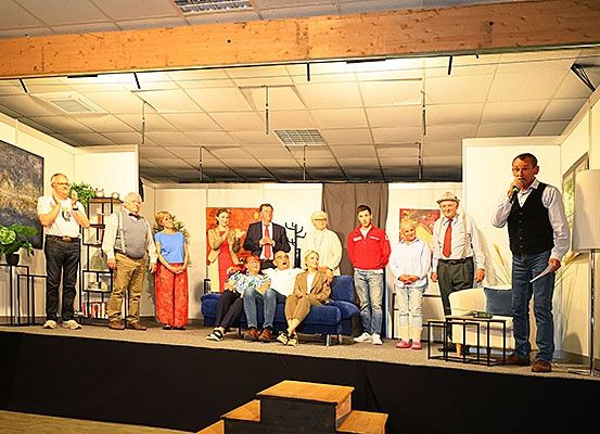 Eine Gruppe von Menschen steht auf einer Bühne, ein Mann spricht in ein Mikrofon, möglicherweise führen sie ein Theaterstück auf.
