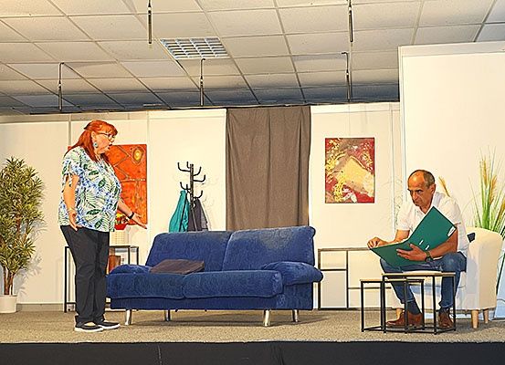Zwei Personen, eine Frau und ein Mann, sind auf einer Bühne mit einer Couch und Stühlen. Die Frau spricht, während der Mann aus einer Mappe liest.