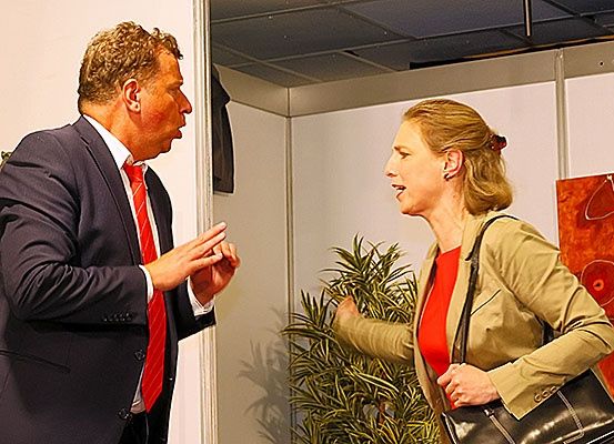 Ein Mann und eine Frau stehen sich gegenüber und führen ein Gespräch. Der Mann trägt Anzug und Krawatte, während die Frau eine Blazerjacke trägt und eine Handtasche hält.