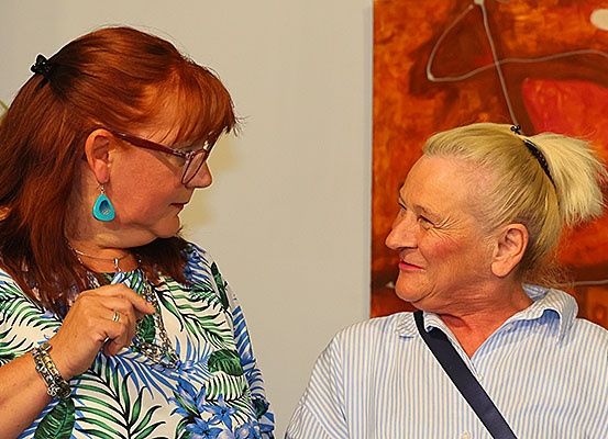 Zwei Frauen stehen vor einer weißen Wand, eine trägt eine Brille und eine Halskette mit einem Anhänger. Die andere Frau hat blonde Haare im Pferdeschwanz. Hinter ihnen befindet sich ein leuchtend orangefarbenes Gemälde.