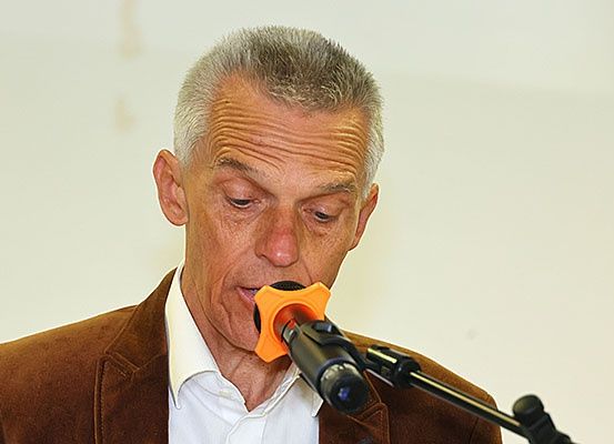 Ein Mann mit grauem Haar spricht in ein Mikrofon. Er trägt eine braune Jacke und ein weißes Hemd. Er sieht konzentriert aus, und sein Mund ist nah am Mikrofon.