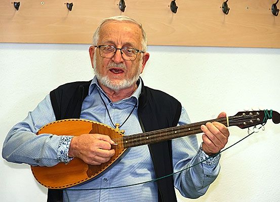 Ein älterer Mann mit Brille spielt eine Laute, während er gegen eine weiße Wand steht. Er trägt ein hellblaues Hemd und eine schwarze Weste.