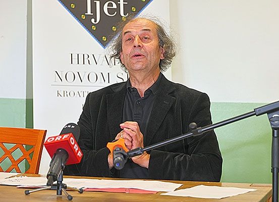 Ein Mann sitzt vor Mikrofonen an einem Schreibtisch, möglicherweise bei einer Pressekonferenz. Er trägt einen schwarzen Mantel. Hinter ihm befindet sich ein Banner mit Text.