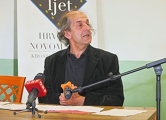 Ein Mann in einem schwarzen Anzug sitzt an einem Tisch mit Mikrofonen, vor einem Banner, das HRV NOVOM steht. Papiere und ein hölzerner Stuhl sind auf dem Tisch.