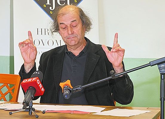 Ein älterer Mann sitzt an einem Schreibtisch und spricht in Mikrofone. Er gestikuliert mit den Fingern. Ein Banner hinter ihm trägt die Aufschrift HRVOM. Papiere und ein Holzstuhl sind auf dem Schreibtisch.