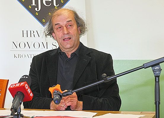 Ein Mann in einem schwarzen Anzug sitzt an einem Schreibtisch mit Papieren und spricht in ein Mikrofon. Hinter ihm steht ein Banner mit der Aufschrift HRVAT NOVOS und KROATIJA.