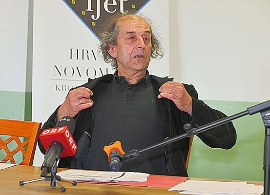 Ein Mann spricht in ein Mikrofon an einem Tisch. Hinter ihm zeigt eine Wand ein Schild mit der Aufschrift 'Hrv Nov'.