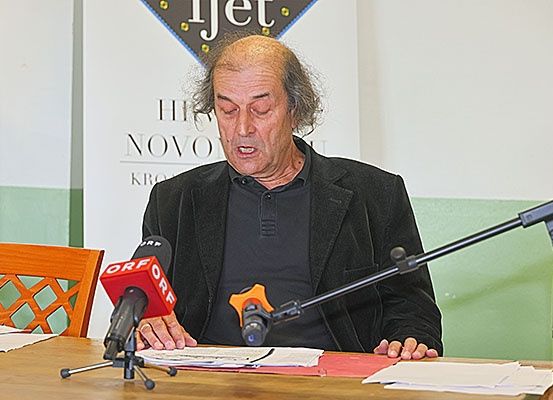 Ein älterer Mann sitzt an einem Tisch mit Mikrofonen und liest von Papieren. Hinter ihm befindet sich ein Banner mit dem Text 'HI NOVO KRO'.