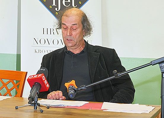 Ein älterer Mann mit grauem Haar sitzt an einem Tisch und spricht in ein Mikrofon. Hinter ihm befindet sich eine Wand mit einem Schild, auf dem HRV NOVO KROATE steht.