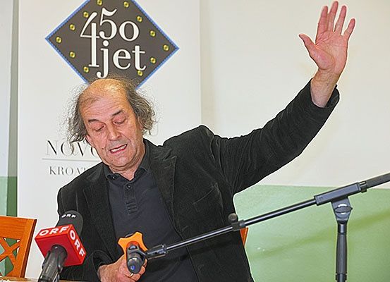Ein älterer Mann mit grauem Haar und einem schwarzen Anzug winkt mit der Hand an einem Podium mit Mikrofonen vor ihm. Hinter ihm ist eine Wand mit einem rautenförmigen Schild, das '450 fjet' steht.