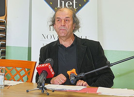 Ein älterer Mann mit grauem Haar spricht in ein Mikrofon an einem Tisch. Papiere und ein Glas Wasser sind auf dem Tisch. Ein Banner mit dem Wort 'liet' ist hinter ihm.