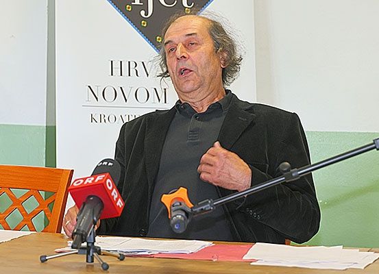 Ein älterer Mann sitzt an einem Schreibtisch und hält eine Rede. Zwei Mikrofone stehen vor ihm, und es gibt Papiere auf dem Tisch. Hinter ihm befindet sich ein weißer Banner mit der Aufschrift 'HRV NOVOM KROATE'.