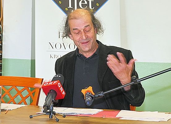 Ein Mann sitzt an einem Schreibtisch mit Mikrofonen vor ihm, scheinbar im Begriff, eine Rede oder ein Interview zu geben. Papiere liegen auf dem Tisch und er trägt eine schwarze Jacke. Hinter ihm ist eine Wand mit einem Schild, das 'Novo Kro' steht.
