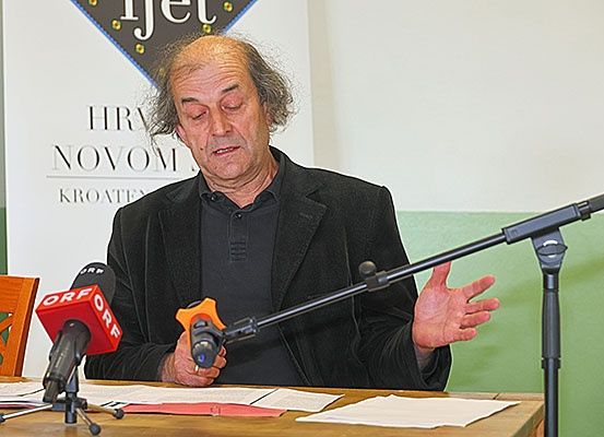 Ein Mann sitzt an einem Schreibtisch und spricht in ein Mikrofon, vor ihm liegen Papiere. Hinter ihm befindet sich ein Banner mit der Aufschrift 'HR NOVOM KROAT'.