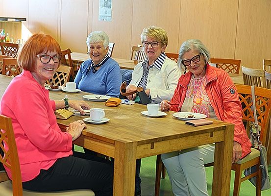 Vier ältere Frauen sitzen an einem Tisch in einem Essbereich, lächeln und schauen zur Kamera. Der Tisch hat Teller, Tassen und verschiedene Gegenstände. Der Raum hat Holzwände und Stühle.