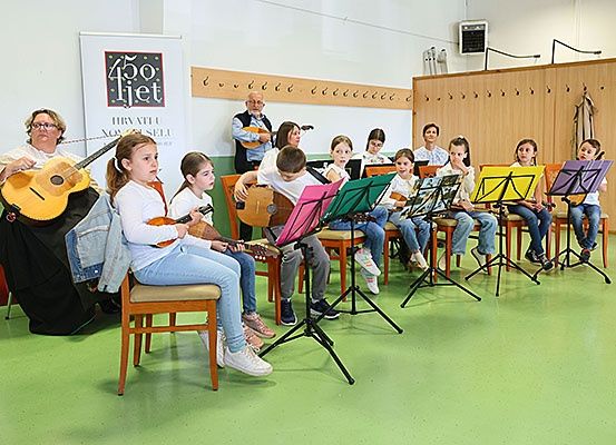 Eine Gruppe von Kindern übt Musik in einem Klassenzimmer. Sie sitzen auf Stühlen mit Notenständern und halten verschiedene Musikinstrumente. Ein Erwachsener steht und hält eine Gitarre.