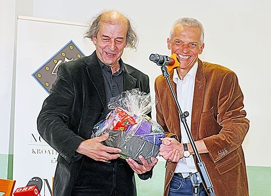 Zwei Männer stehen vor einem Mikrofon, einer hält ein in Plastik eingewickeltes Geschenk, und beide lächeln in die Kamera. Hinter ihnen steht ein Schild mit dem Wort 'Kroatien'.