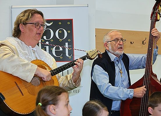 Zwei Männer spielen Gitarre und Bass vor einer Gruppe von Kindern, wahrscheinlich in einem Klassenzimmer.