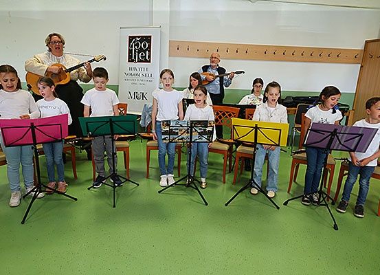 Eine Gruppe von Kindern und Erwachsenen versammelt sich in einem Raum und übt Musik. Sie halten Notenständer und singen. Zwei Erwachsene spielen Instrumente.
