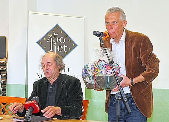 Zwei Männer stehen vor einem Mikrofon, einer hält einen Geschenkkorb. Hinter ihnen steht ein Schild mit der Aufschrift '450 Let' auf weißem Hintergrund. Der sitzende Mann hält ein Mikrofon und scheint zu sprechen.