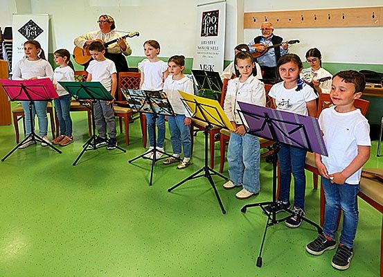 Eine Gruppe von Kindern mit Musikständern in einem Raum. Einige halten Gitarren und andere stehen hinter ihnen. Zwei Erwachsene spielen Gitarren im Hintergrund.