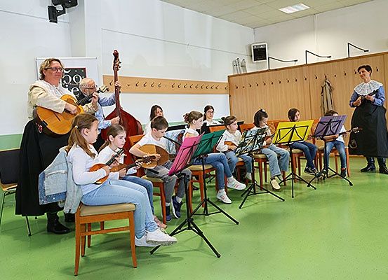 Eine Gruppe von Kindern sitzt in einem Klassenzimmer mit Musikinstrumenten. Eine Frau steht in der Nähe, und eine andere Frau steht im Hintergrund.