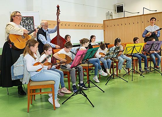 Eine Gruppe von Kindern und Erwachsenen spielt Musikinstrumente in einem Klassenzimmer. Zwei Erwachsene stehen und halten Gitarren. Die Kinder sitzen auf Stühlen mit Notenständern.