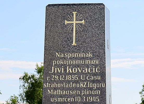 Ein Gedenkstein mit einem goldenen Kreuz oben. Es steht: 'Zum Gedenken an den verstorbenen Jivi Kovacic, geboren am 29. Dezember 1895, gestorben in Mauthausen am 10. März 1945.' Bäume und ein blauer Himmel im Hintergrund.