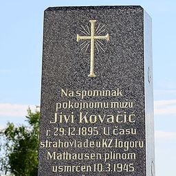Ein Gedenkstein mit einem goldenen Kreuz oben. Es steht: 'Zum Gedenken an den verstorbenen Jivi Kovacic, geboren am 29. Dezember 1895, gestorben in Mauthausen am 10. März 1945.' Bäume und ein blauer Himmel im Hintergrund.