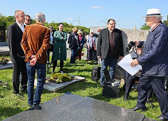 Mehrere Menschen stehen auf einem Friedhof, einer hält ein Papier und spricht. Ein anderer hält eine Kamera und macht ein Foto. Hinter ihnen steht ein Grabstein mit einer Pflanze, und in der Ferne sind Windmühlen zu sehen.