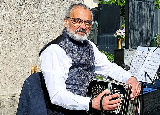 Ein Mann mit Brille und Bart spielt Akkordeon auf einem Friedhof mit Blumen im Hintergrund.