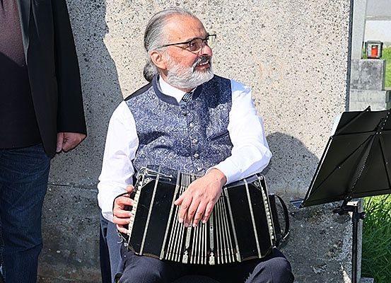 Ein älterer Mann mit Brille und blauer Weste spielt Akkordeon, während er gegen eine Wand gelehnt sitzt, ein anderer Mann in Anzug steht hinter ihm.